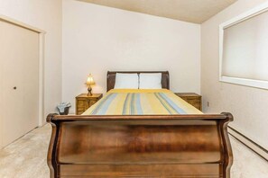 A bedroom with a wooden bed, a nightstand, a lamp, and a window with blinds.