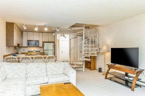 Ein gemütlicher Wohnraum mit Sofa, Esstisch, Fernseher auf einem Schrank, Küchenzeile und Wendeltreppe.