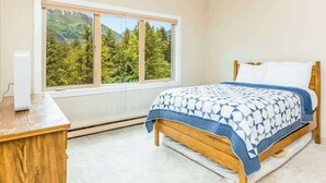 Een slaapkamer met een bed, een houten nachtkastje, een raam met uitzicht op bomen en bergen, en een witte muur.