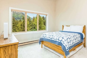 Ein Schlafzimmer mit einem Bett, einem Nachttisch aus Holz, einem Fenster mit Blick auf Bäume und Berge und einer weißen Wand.