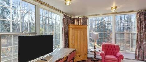 Um quarto com uma grande janela, uma mesa de madeira, uma televisão, uma cadeira vermelha e um armário.