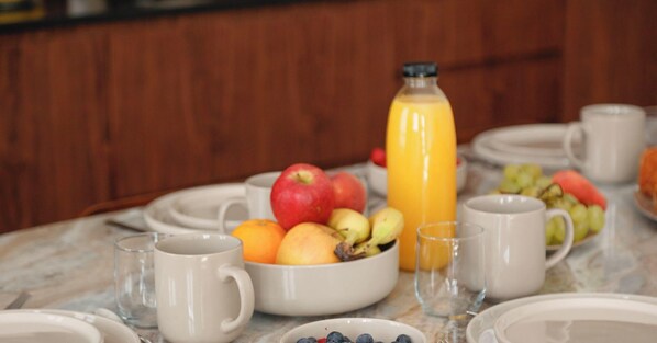 Una mesa de mármol con un bol de frutas, una botella de jugo y tazas blancas.