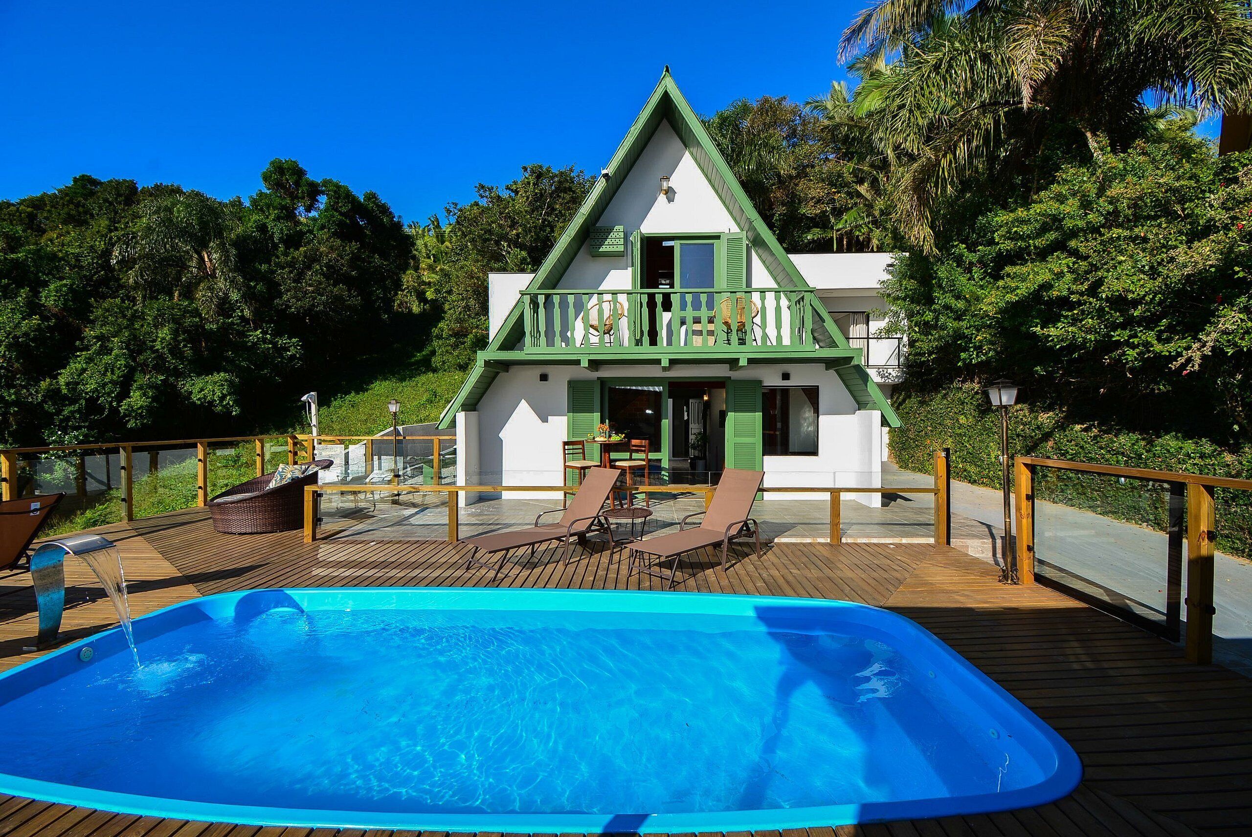 A pool with lounge chairs, a wooden deck, and a house with a green roof and balcony.