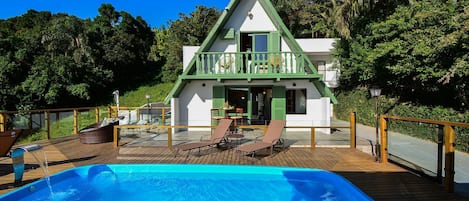 A pool with lounge chairs, a wooden deck, and a house with a green roof and balcony.