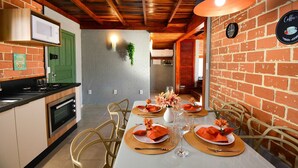 Un comedor con pared de ladrillo, techo de madera y una mesa preparada para cuatro, con servilletas anaranjadas y un florero con flores.