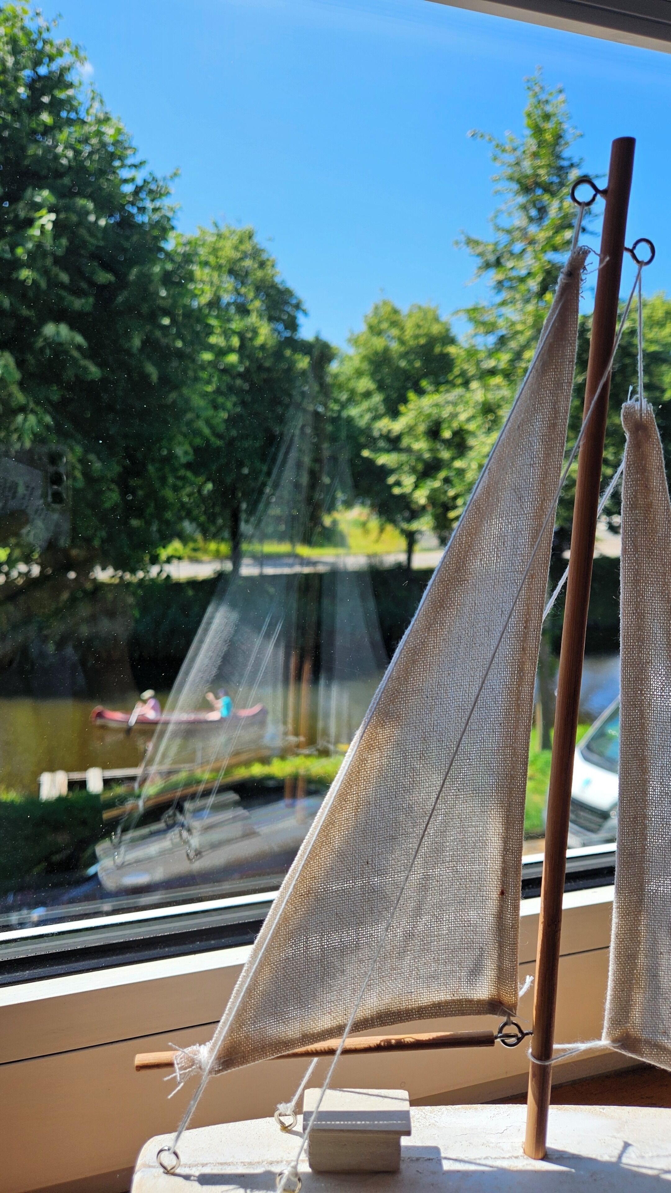 Un modelo de velero con vela de red y mástil de madera, situado en un interior con vista a un río y follaje exterior.
