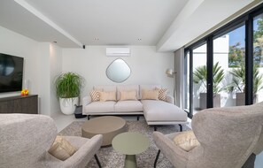 Ein modernes Wohnzimmer mit einem Ecksofa, zwei Sesseln, einem Couchtisch, einem großen Spiegel und einem Fernseher auf einem Standfuß.