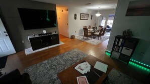 Ein Wohnzimmer mit einem Flachbildfernseher, einem Couchtisch mit Spielkarten, einem Essbereich mit Stühlen und einem Bücherregal mit Dekoration.