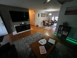 Ein Wohnzimmer mit einem Flachbildfernseher, einem Couchtisch mit Spielkarten, einem Essbereich mit Stühlen und einem Bücherregal mit Dekoration.