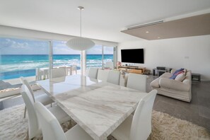 Ett modernt vardagsrum med ett stort fönster med utsikt över havet, en matplats med ett bord i marmor och en mysig sittgrupp med en soffa och en platt-TV.