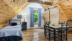 Ein gemütliches Schlafzimmer im Dachgeschoss mit Holzbalkendecke, einem Bett, einem Tisch mit Stühlen und einem Fenster mit Vorhängen.