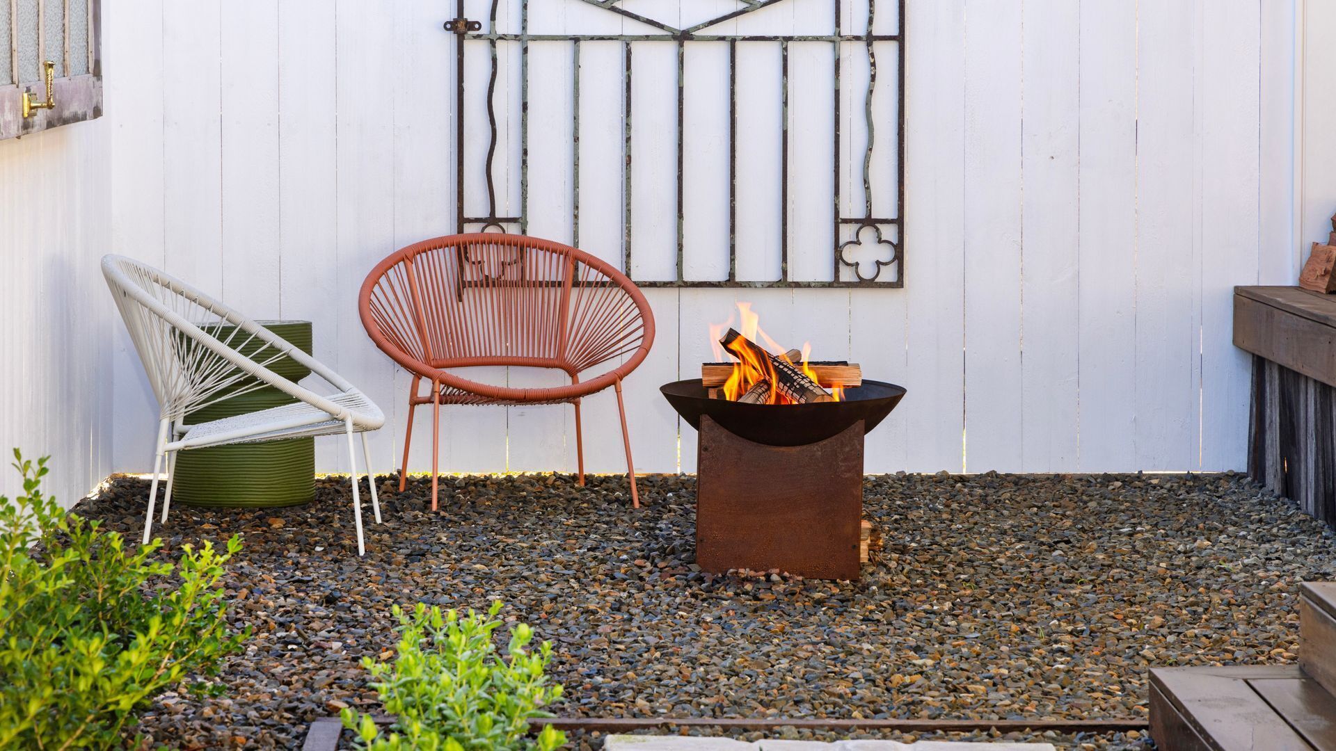 A patio with two chairs, a fire pit, and a white wall with a metal gate.