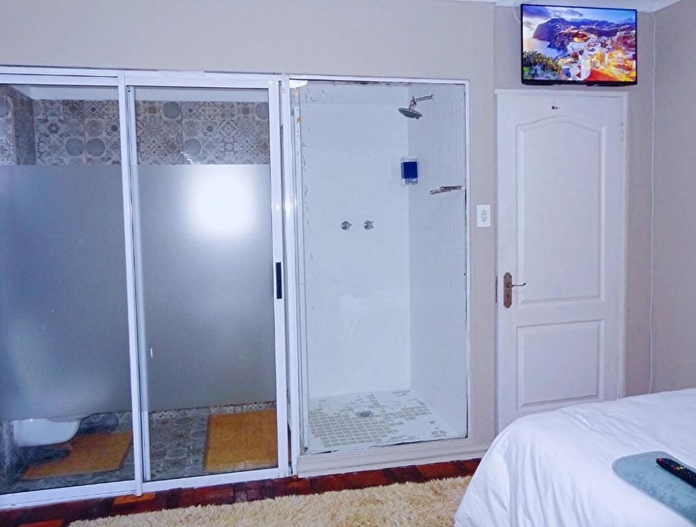 A room with a bed, a white door, a shower area with glass doors, and a television on the wall.