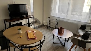 A living room with a TV, two dining tables, chairs, a plant, and a window with curtains. - Quiet and convenient apartment (Évaux-les-Bains)