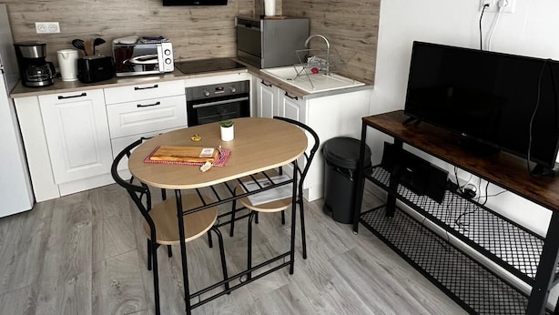 A modern kitchen with white cabinets, a wooden countertop, a dining table with chairs, a TV on a stand, and a black trash bin. - Quiet and convenient apartment (Évaux-les-Bains)