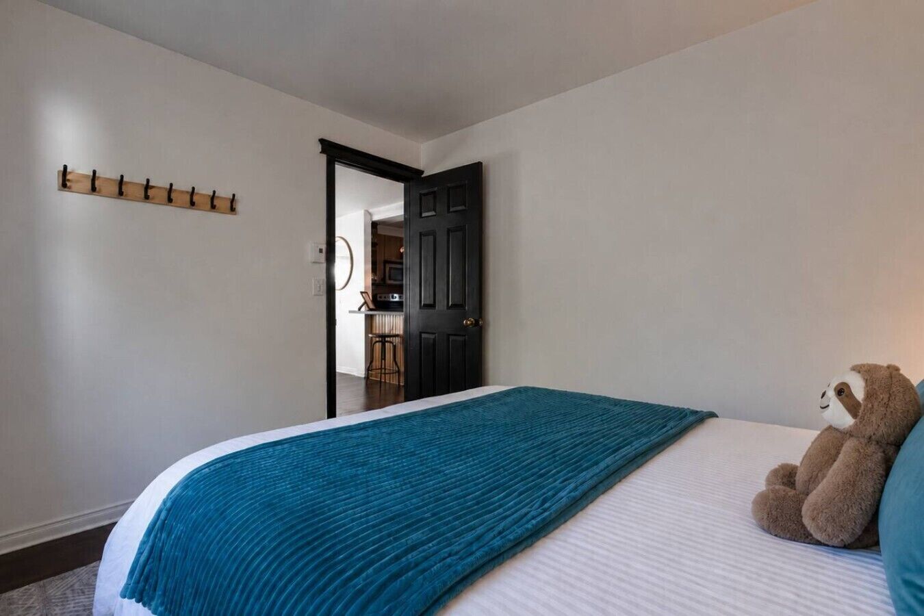 Een slaapkamer met een bed, een pluche luiaard, een blauwgroene deken en een houten kapstok.