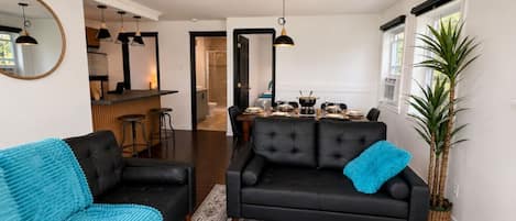 Una sala moderna con un sofá negro, un sillón azul, un comedor con mesa dispuesta para cuatro personas y una cocina al fondo.