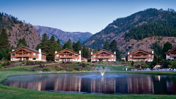 A row of wooden cabins with balconies, a pond with a fountain, and mountains in the background. - Leavenworth 2BR Mountain Villa – Full Kitchen, Balcony & Near Bavarian Village (Leavenworth)