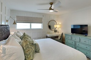 Une chambre à coucher comprenant un lit, un bureau, une commode, un téléviseur, un ventilateur de plafond et une fenêtre avec des stores.