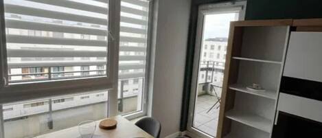 Una habitación moderna con una mesa de madera, sillas negras, una estantería blanca y ventanas amplias con persianas.