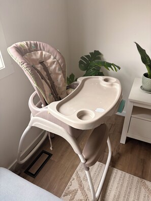 A high chair with a cushioned seat and backrest, positioned in a room with a wooden floor, a white wall, and a potted plant. - Luxury 4BR Calgary Home|Families| Sleeps 8|AC& Free Parking & WiFi|Near Airport| (Calgary)