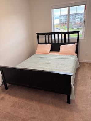 Un lit simple avec une tête de lit, deux oreillers et une couvre-lit matelassée, dans une pièce dont le sol est recouvert de moquette et qui possède une fenêtre donnant sur des immeubles.