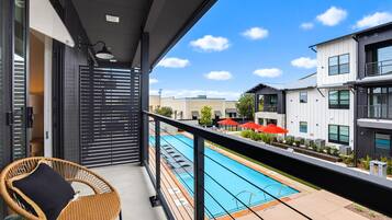 A balcony with a wicker chair, a pool, and modern buildings in the background.