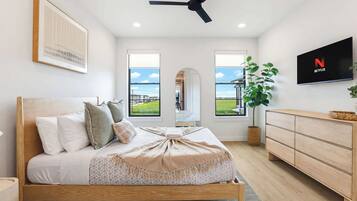A bedroom with a bed, a TV, a ceiling fan, a dresser, and large windows.