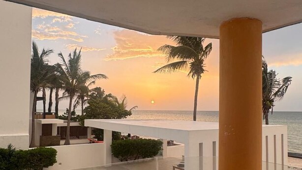 Een balkon met uitzicht op de ondergaande zon boven de oceaan, palmbomen en een zwembad.