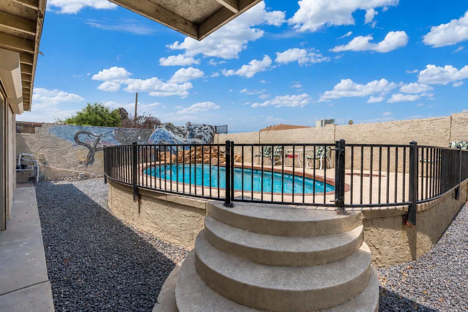 Une piscine entourée d’une clôture en métal noir, avec un escalier en pierre et un ciel d’un bleu limpide.