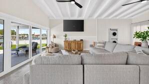 Ein modernes Wohnzimmer mit einem großen, grauen Ecksofa, einem Holzkasten, einem Flachbildfernseher, Deckenventilatoren und Blick durch Glastüren auf einen Außenbereich.