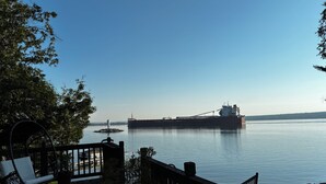 Ein großes Schiff liegt in ruhigem Wasser vor Anker, ein Holterrasse mit Stuhl und Pflanztopf, und ein klarer Himmel.