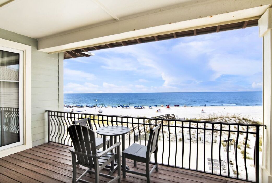 Een houten terras met twee stoelen en een tafel, met uitzicht op een strand met mensen en parasols.