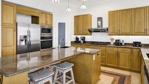 A modern kitchen with wooden cabinets, stainless steel appliances, a granite countertop, and bar stools. - Dixie Tide - Hammock Dunes West (Orange Beach)