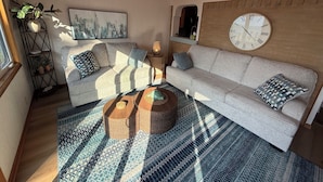 Una sala de estar acogedora con un sofá, una mesa redonda para café, un reloj de pared grande y un cuadro con una vista urbana.