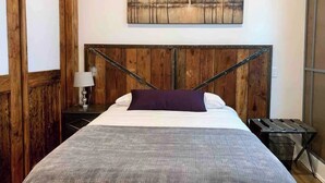 Et soveværelse med sengegavl i træ, en seng med gråt tæppe, et natbord med lampe og et maleri på væggen.