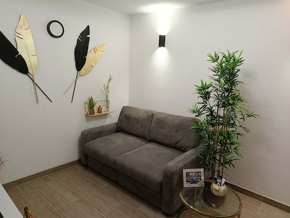 Una sala de estar con un sofá gris, una mesa de centro de vidrio, un aplique de pared, plumas decorativas y una planta en maceta.