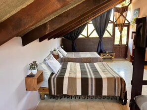Ein Schlafzimmer mit zwei Betten, einer Holzbalkendecke, einem Fenster mit Vorhängen und einem kleinen Nachttisch mit einer Pflanze.