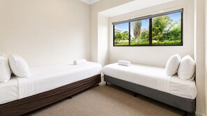 A room with two single beds, a window with a view of greenery, and a carpeted floor. - Anglesea Family Bungalow Pool View by Book Jervis Bay (Huskisson)