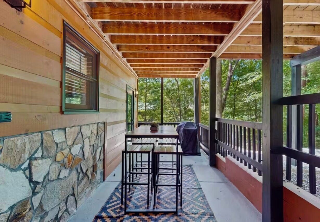 A covered porch with a stone wall, a wooden table with chairs, and a grill.