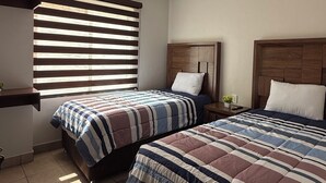 Ein Schlafzimmer mit zwei Betten, einem Fenster mit Jalousien, einer Deckenleuchte und einem kleinen Nachttisch mit einer Pflanze.
