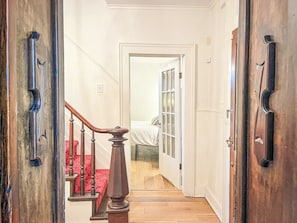 A hallway with a wooden staircase, a door leading to a room with a bed, and a window.