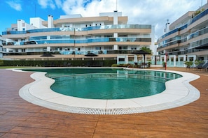 A swimming pool with a wavy edge, surrounded by a wooden deck, and a modern apartment building in the background. - Luxury Penthouse in Jurerê Internacional: 150m² Terrace, Private Pool and Suite (Florianópolis)