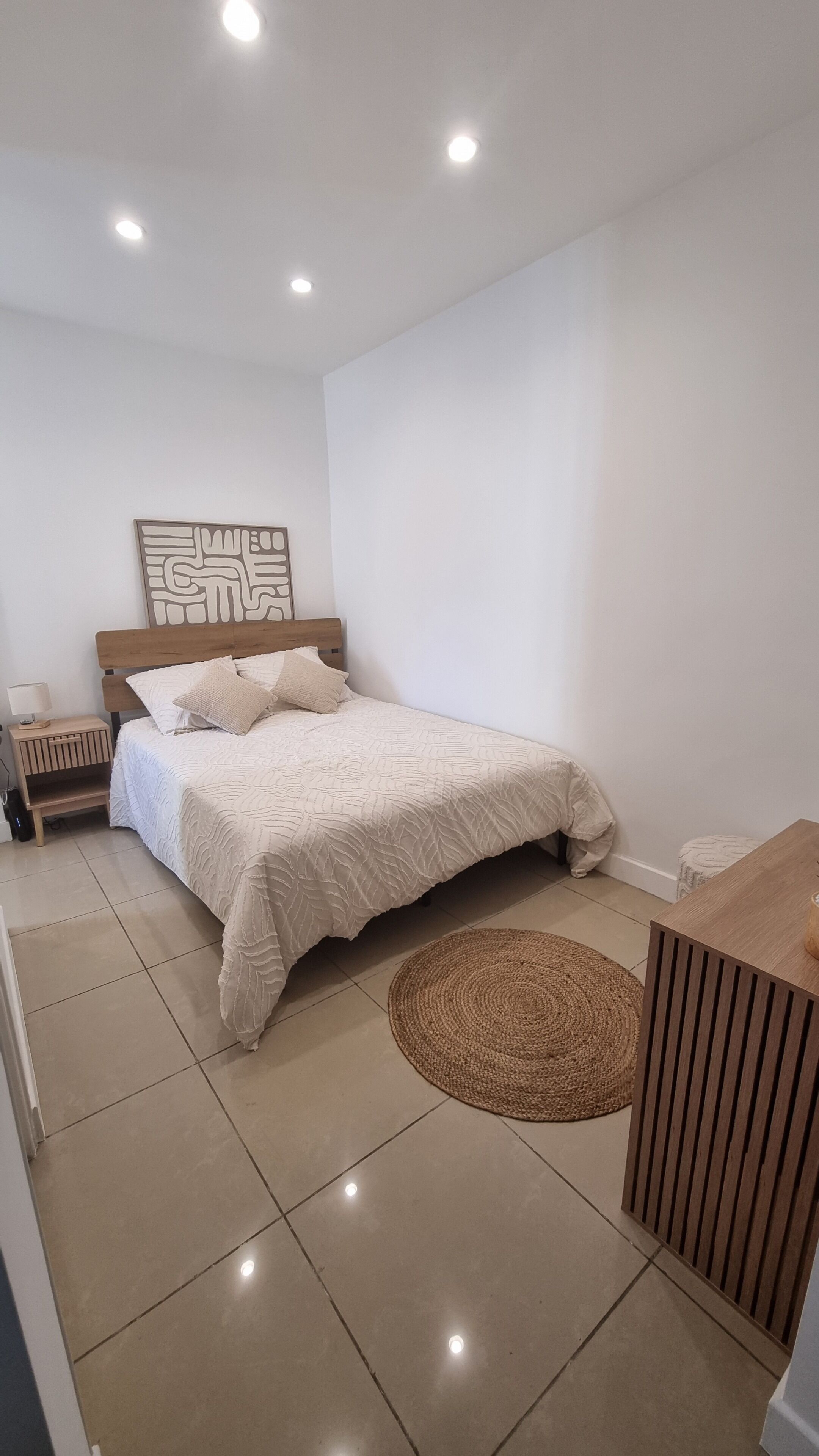 Un dormitorio con cama, mesita de noche, una obra de arte en la pared, alfombra circular tejida y un armario de madera.