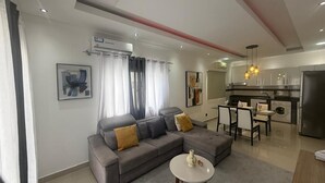 A modern living room with a grey sofa, a small round table, and a kitchen area with a dining table and chairs.