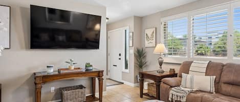 A living room with a flat-screen TV, a brown leather sofa, a coffee table, and a large window with blinds.