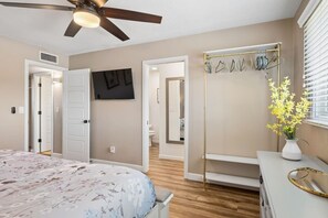 Une chambre à coucher comprenant un lit, un ventilateur de plafond, une télévision, une coiffeuse avec un miroir et un couloir menant à la salle de bain.