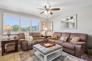 A living room with a brown leather sofa, a white ottoman, a ceiling fan, and a large window with blinds. - Old Town Scottsdale - Pool - Grill - Sun Loungers (Scottsdale)