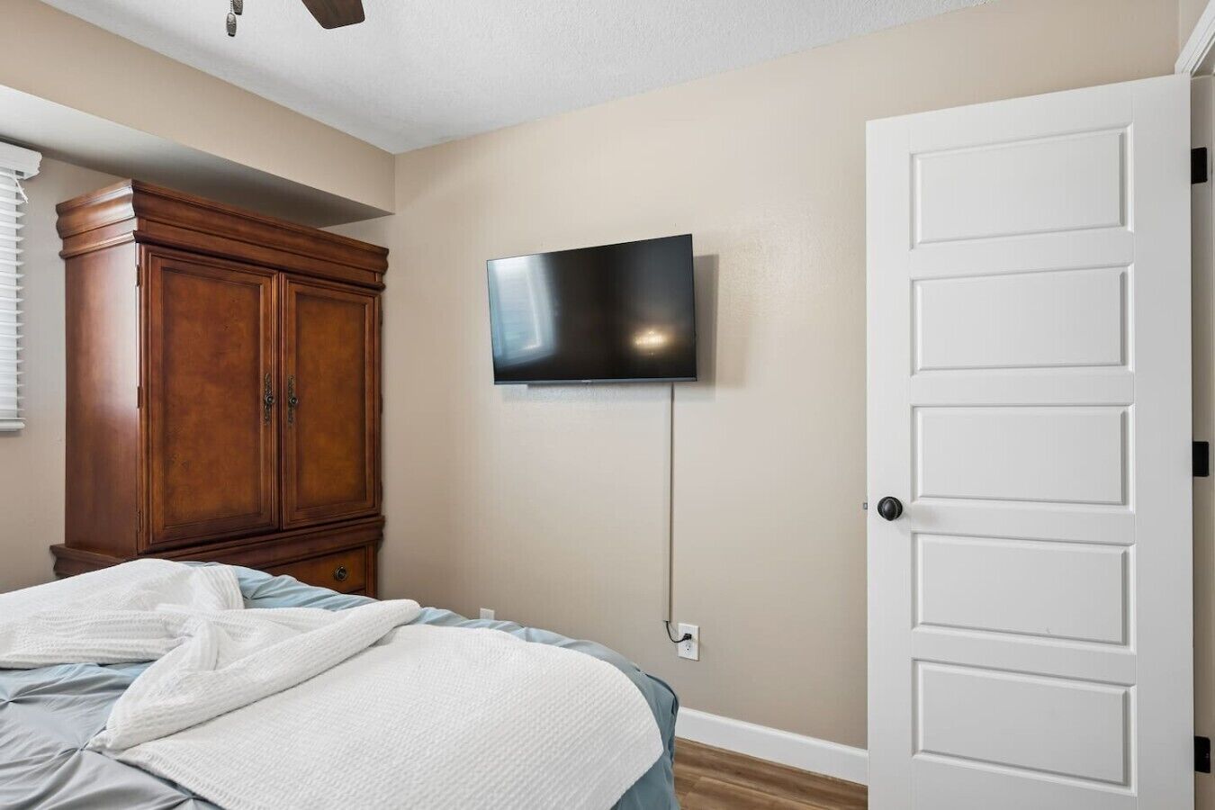 Une chambre à coucher avec un lit, une armoire en bois, un téléviseur fixé au mur et une porte blanche.