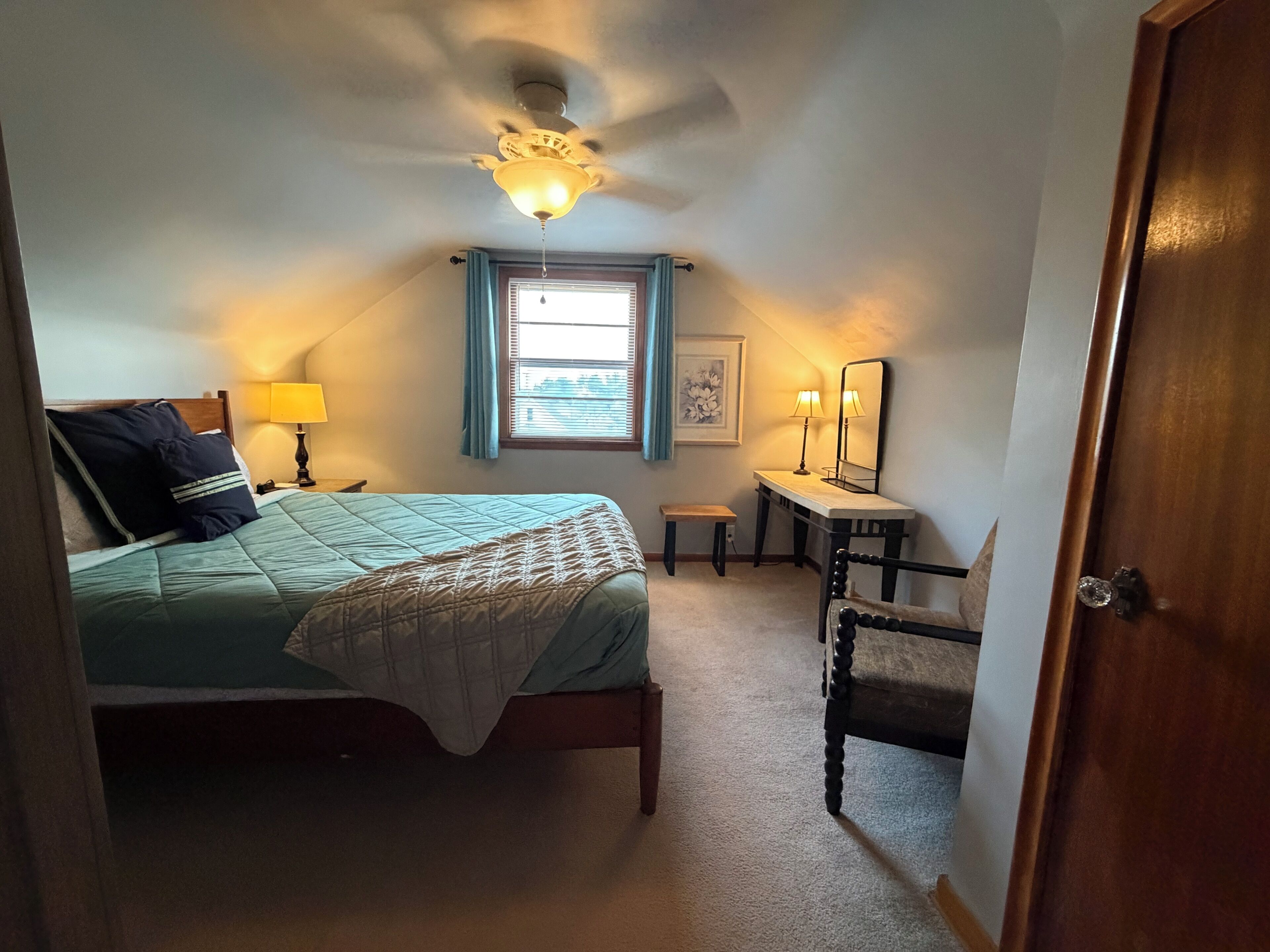 Un dormitorio con cama, mesita de noche, lámpara, ventana, ventilador de techo y puerta.